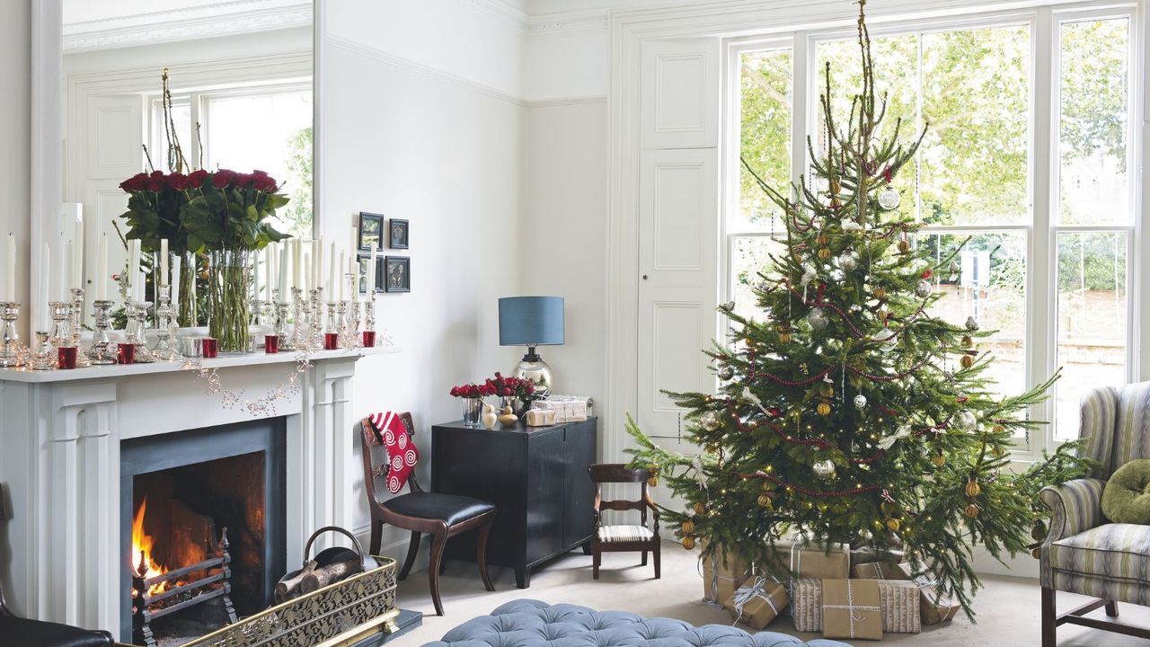 Festive living room with large Christmas tree, padded blue ottoman, white fireplace and mantelpiece with large mirror, white walls and large windows with white shutters.