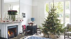 Festive living room with large Christmas tree, padded blue ottoman, white fireplace and mantelpiece with large mirror, white walls and large windows with white shutters.
