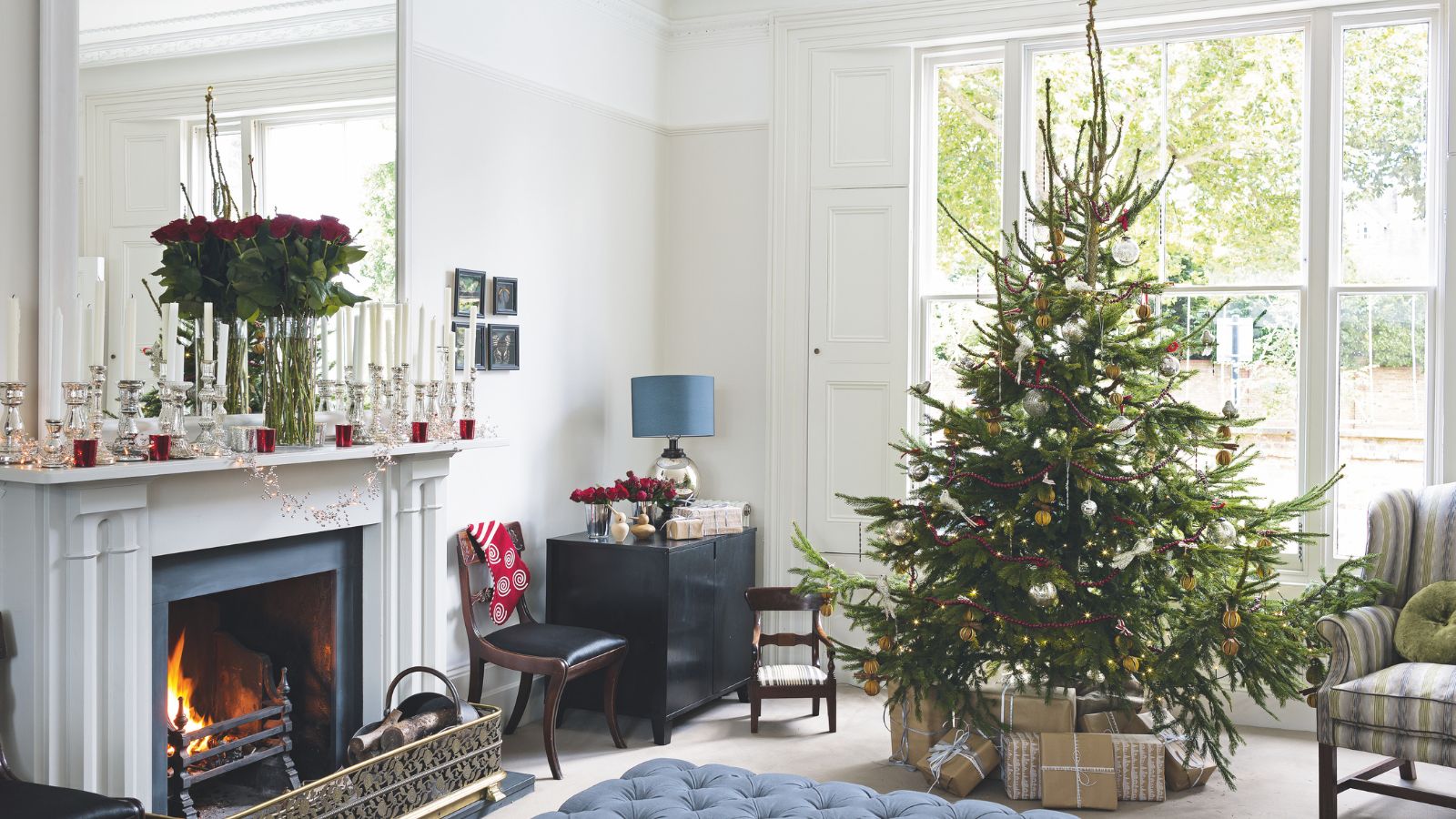 Festive living room with large Christmas tree, padded blue ottoman, white fireplace and mantelpiece with large mirror, white walls and large windows with white shutters.