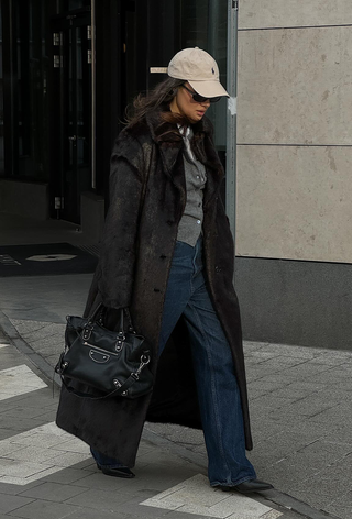 what to wear in 30-degree temperatures shown in a photo of a woman walking down the sidewalk wearing a tan baseball cap with a dark brown fur coat over a gray cardigan and white t-shirt styled with dark wash jeans, black ankle boots, and a black bag