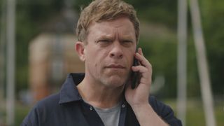 Dylan in navy overalls, standing on his boat, receiving a call about the crash.