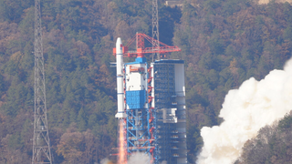 a white rocket launches with a tree-covered hill in the background.