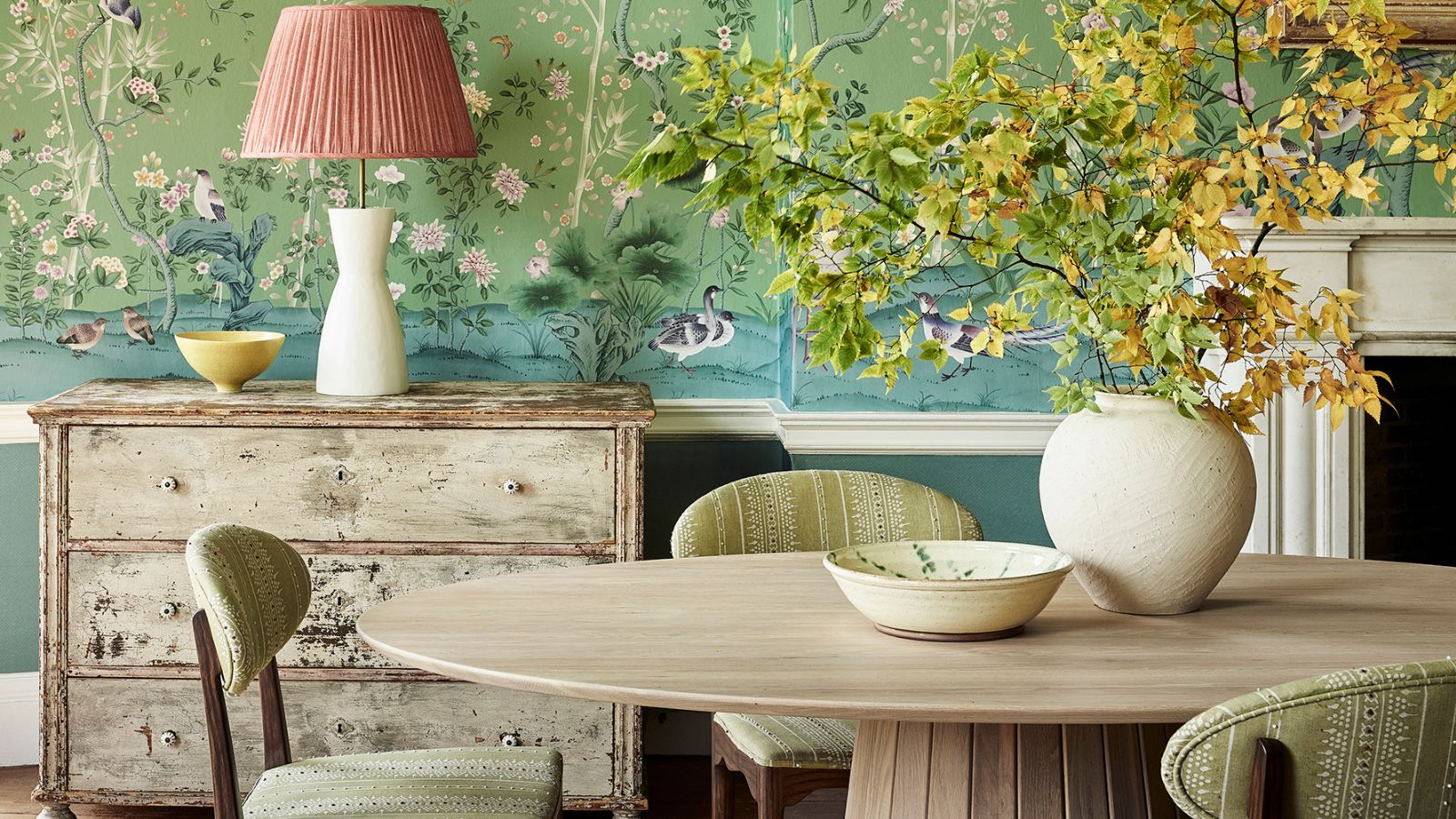 Dining area with Fromental&#039;s green floral wallpaper and round wooden dining table with mid century pale green upholstered dining chairs. There is a distressed sideboard in view with a cream lamp, and pink lampshade