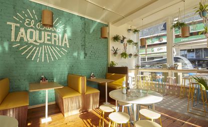 Exposed face-brick wall painted an eye-catching shade of turquoise blue with restaurant&#039;s name on the wall, creamy walls with potted plants, colouful high chairs, wood and yellow leather sofas and tables