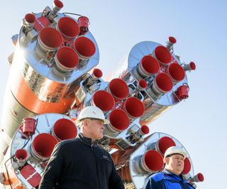 Two people a standing in front of the chrome bottom of a soyuz rocket. the quintuple-cored rocket has four primary engines on each bottom stage.