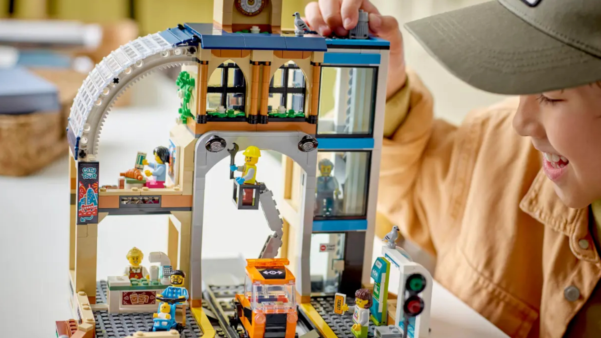 A boy in a cap smiles while placing a piece on top of the Central Train Station Lego set, which sits on top of a white surface