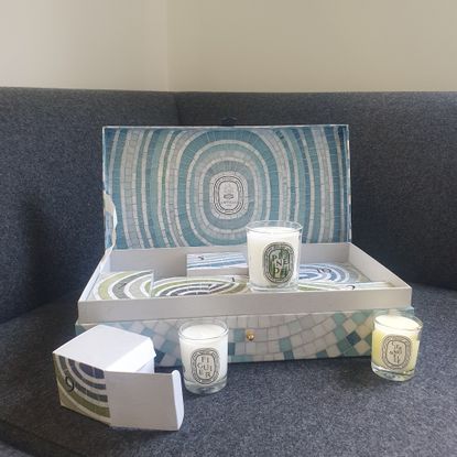 Open Diptyque Summer Essentials calendar on a dark grey sofa with three candles arranged around the box