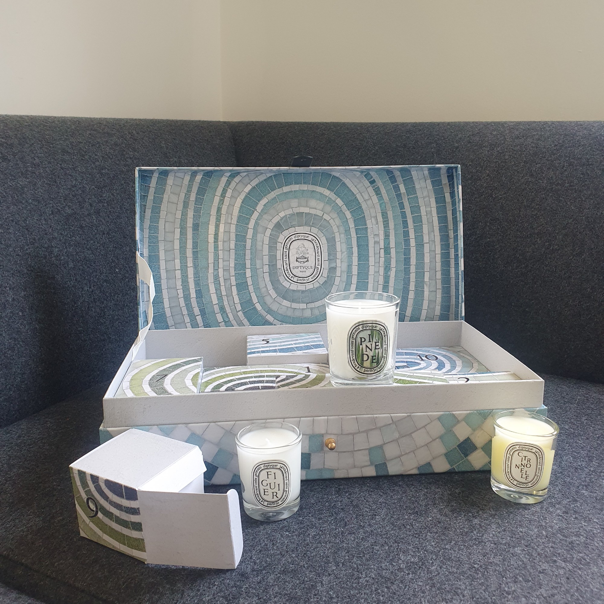 Open Diptyque Summer Essentials calendar on a dark grey sofa with three candles arranged around the box