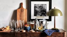 Crate & Barrel x NFL-branded beer glasses, silver barware, and a wooden serving board pictured atop a rustic burlwood credenza alongside a moss-green table lamp and family photo.