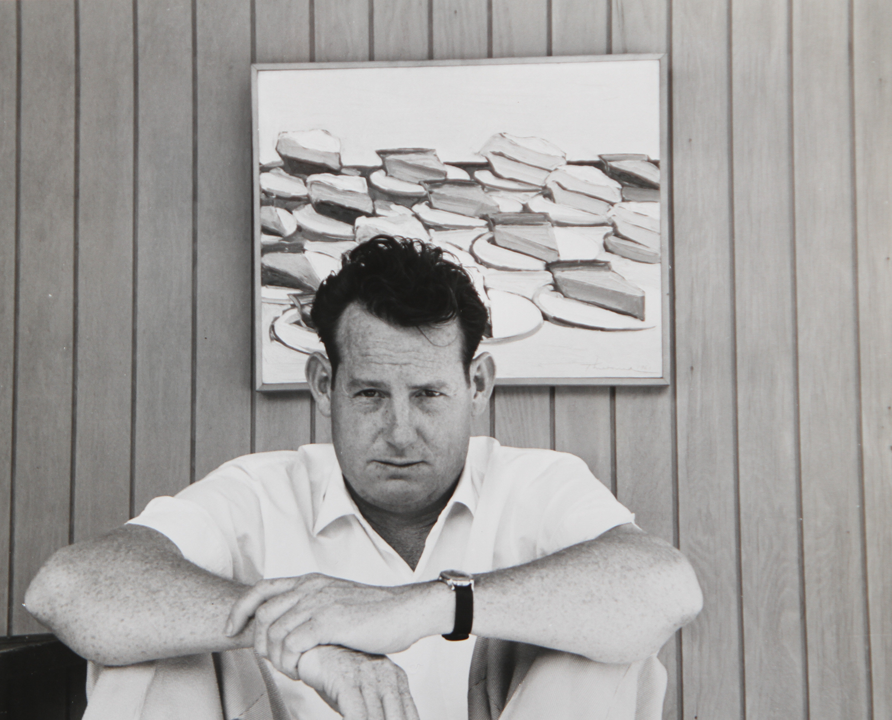Wayne Thiebaud in 1961, sitting with crossed arms infront of one of his still life paintings of slices of cake