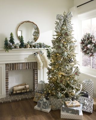 White living room with a fireplace and decorated Christmas mantel. There is a Christmas tree in the corner beside the fireplace that has soft white and velvet decorations. There are silver presents under the tree.