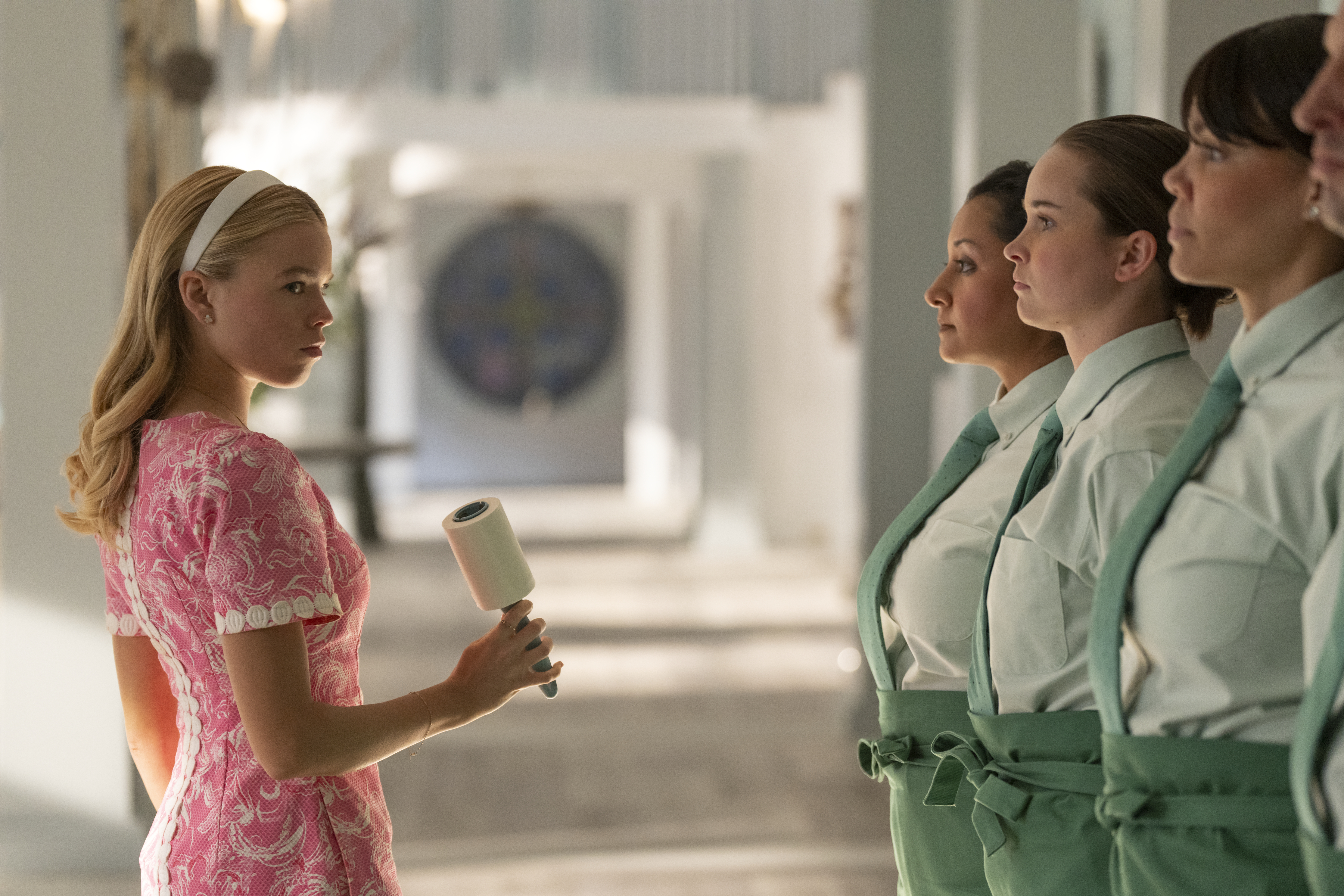 milly alcock as simone wearing a pink dress and holding a lint roller in front of the house help in the tv show sirens