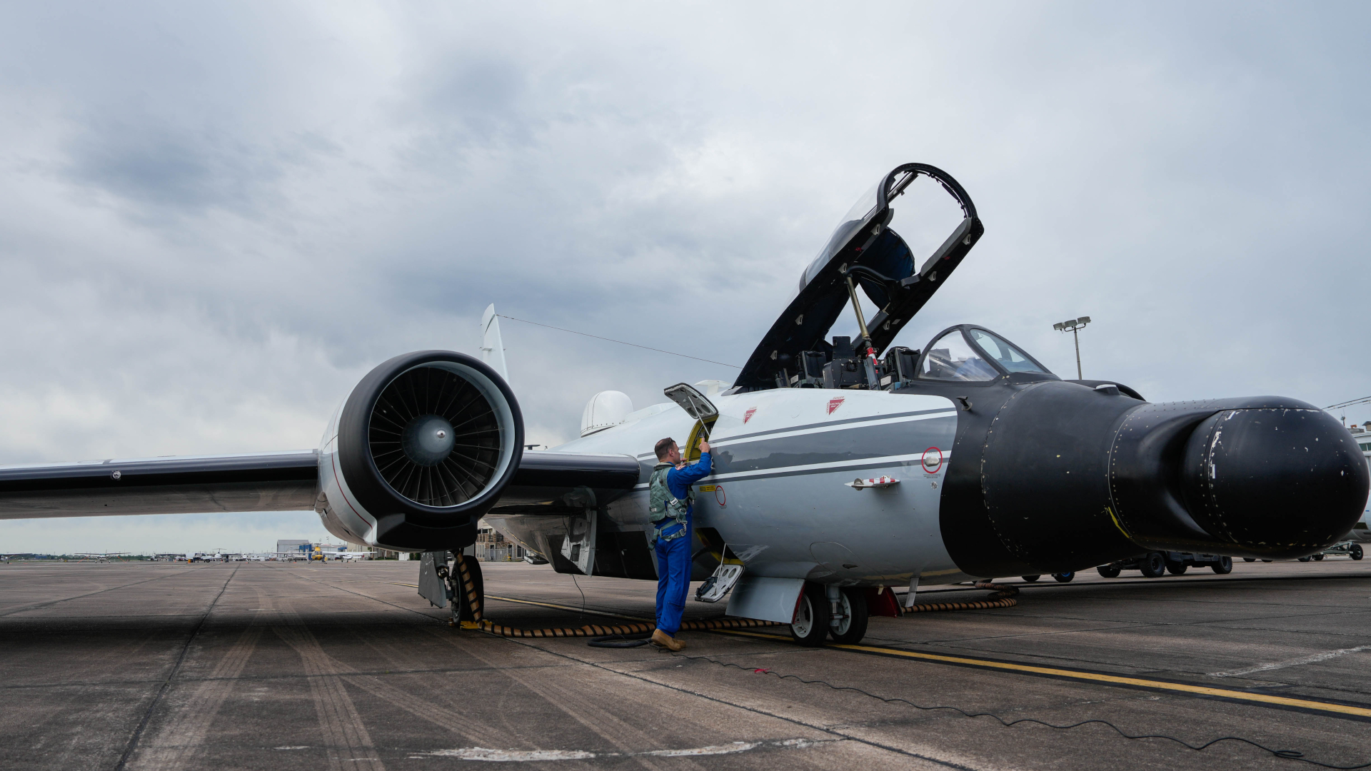 A man wearing a blue flight suit peers into an open compartment on the side of a black and white plane which sits on the runway with its cockpit open