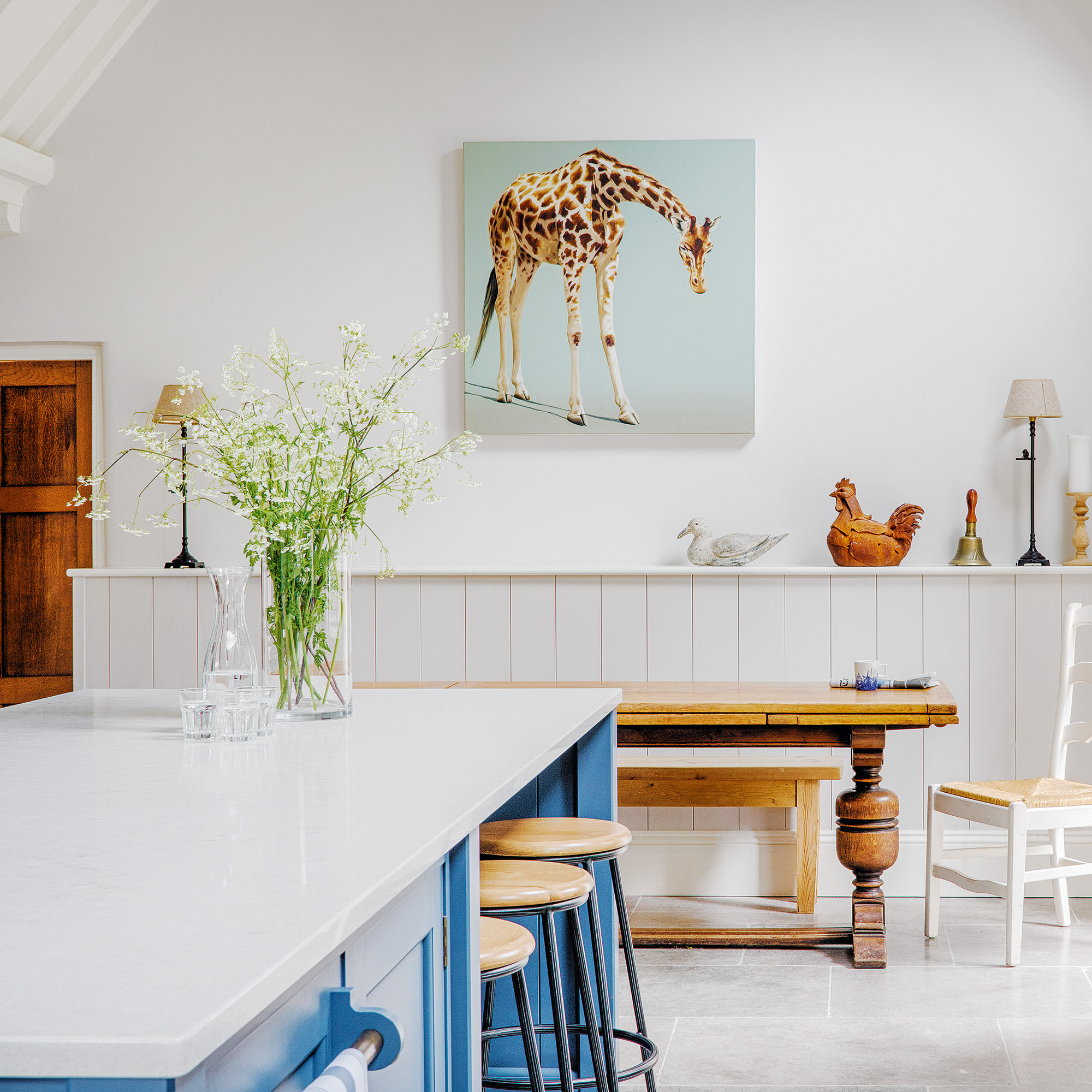 kitchen with white walls, wooden table and bench against the back wall and kitchen island with quartz worktop and blue painted base on back wall is an illustrated painting of a giraffe on a pale blue background