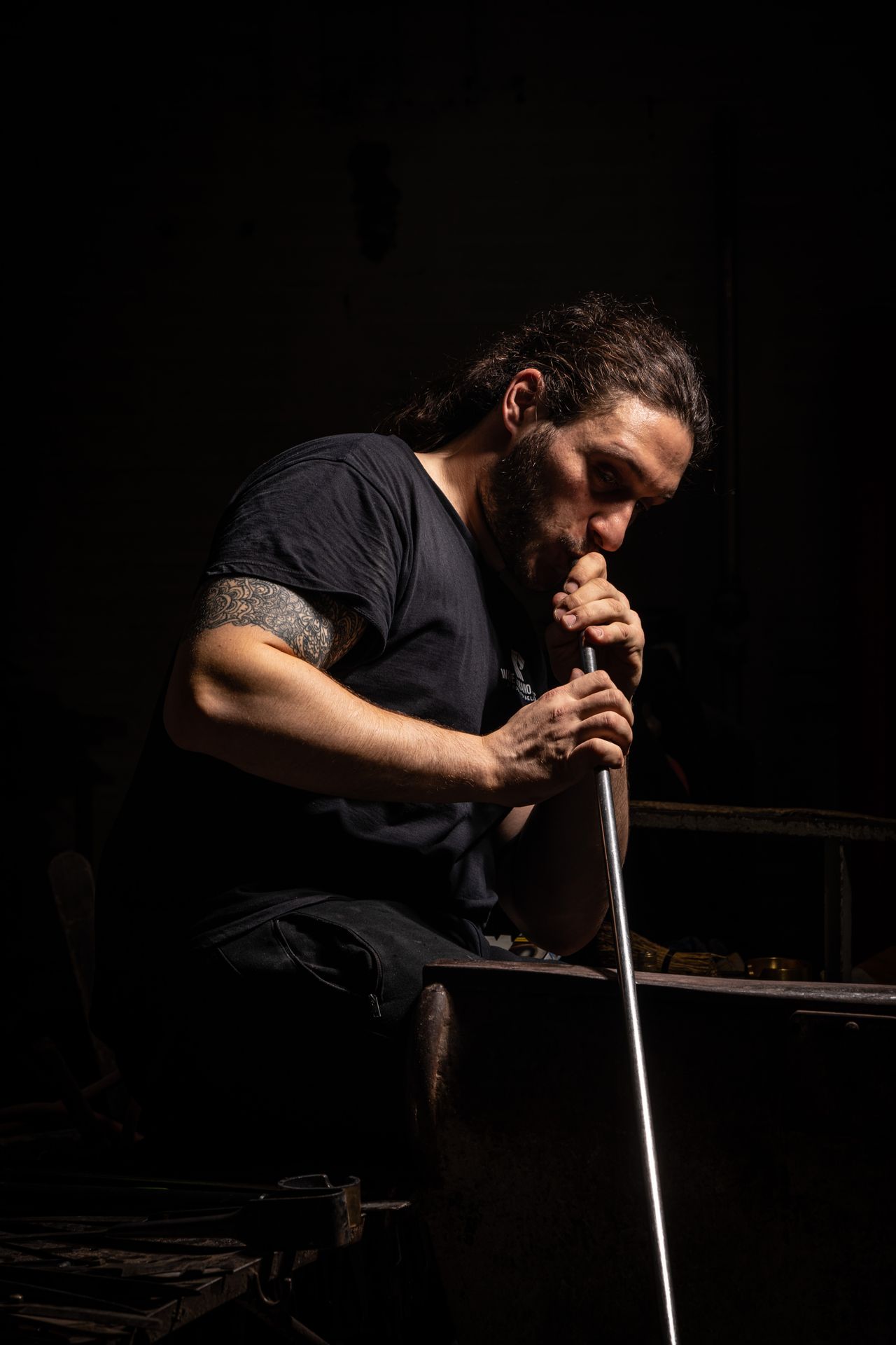 A young man dressed in black clothes blows glass in a darkly lit glass furnace.