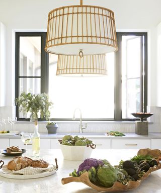A white kitchen with a countertop with a wooden tray of artichokes and beetroot, a white tray with bread on it, a white basket with artichokes in it, a bottle of white wine and a glass of it, with a rattan pendant light above it and a black window beyond it