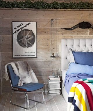 bedroom with lime-washed boards on both the floor and walls, a grey bed with blue coves and colourful striped throw, a picture on the wall next to a hanging lamp above a pile of magazines, a blue chair with a sheepskin draped over it, and a green foliage garland along the top of the wall