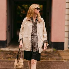 A woman wears a black skirt, plaid shirt, barn jacket, black loafers, and a handbag while posing for a photo. 