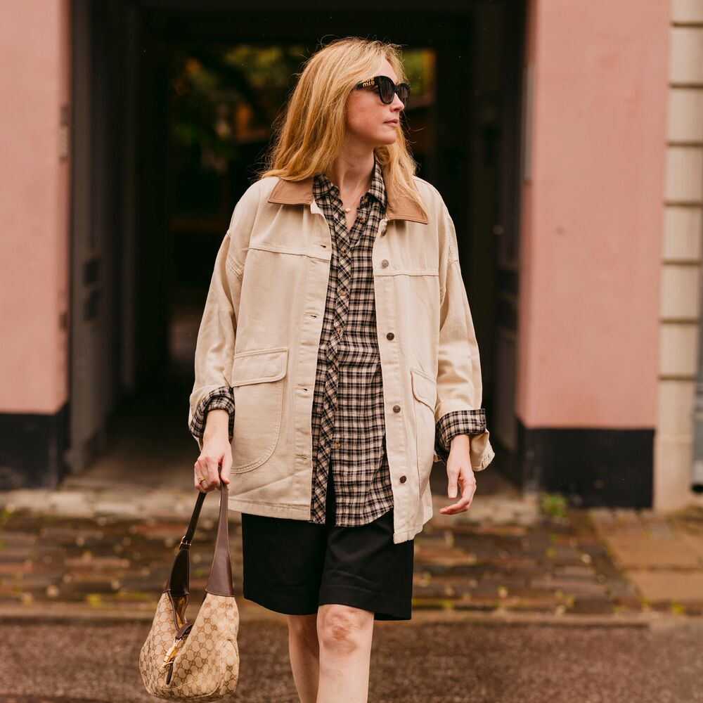 A woman wears a black skirt, plaid shirt, barn jacket, black loafers, and a handbag while posing for a photo. 
