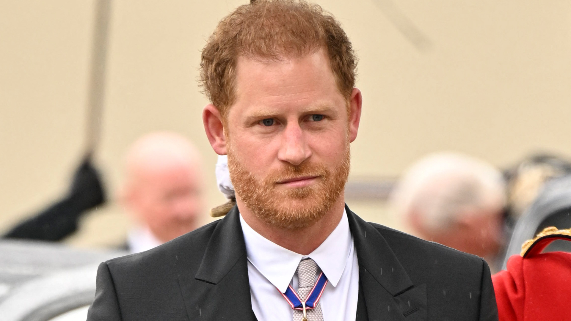 Prince Harry is a man with ginger hair, and a slight smirk on his face, wearing a black suit with military medals attached to the lapel