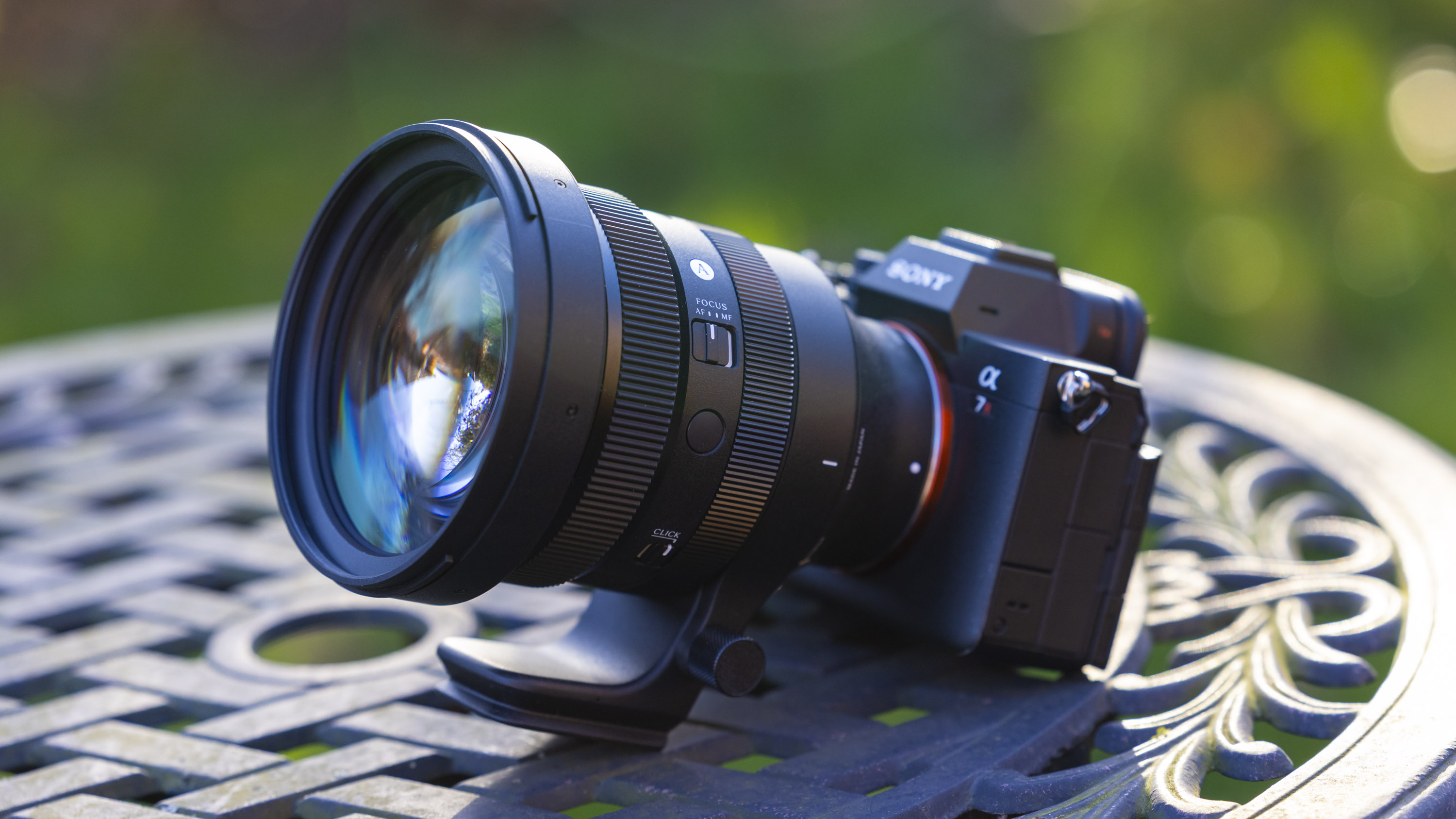 The Sigma 135mm f/1.4 DG | Art lens attached to a Sony A7R IV body positioned outdoors on a sunny day on a metal outdoor table with a lattice pattern