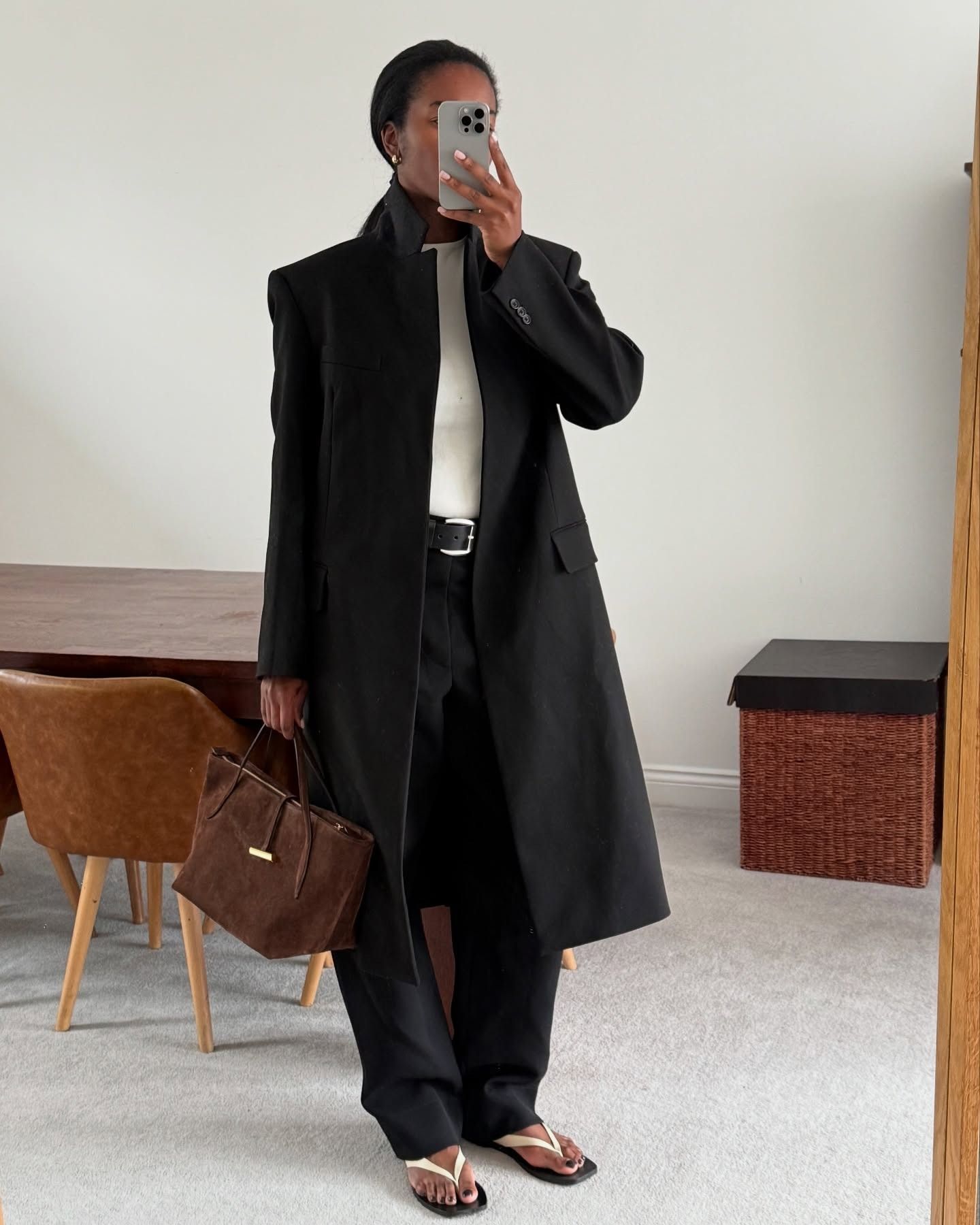 Marilyn wears a black long jacket, white tee, black pants, white sandals, and a brown suede bag.