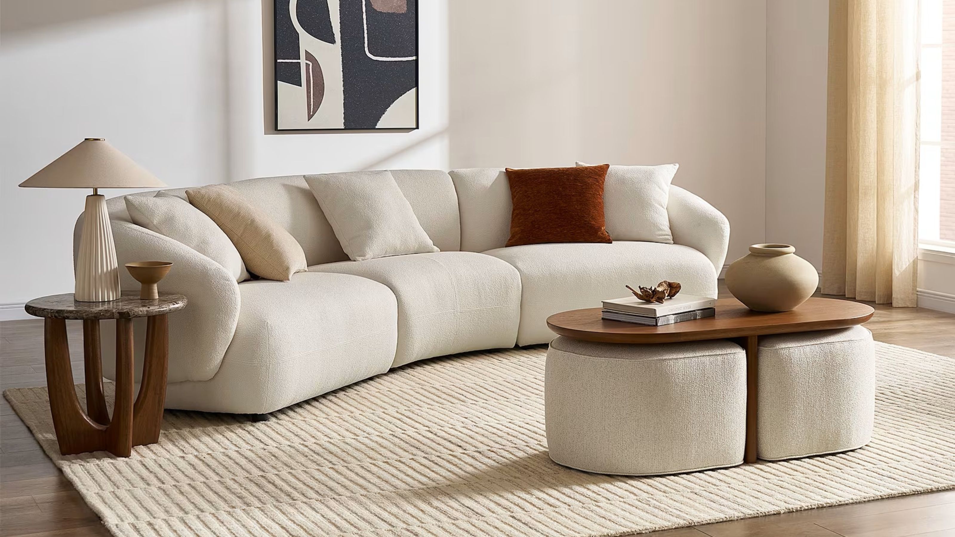 Image of a white living room with a large white, curved sofa that has a white rug underneath and a nesting coffee table and stool set in front of it.