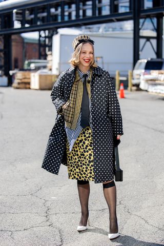 Chloe King wears beige black striped hat, black jacket with dots print, animal yellow print skirt, black tie, checkered blazer, knee high tights outside Brandon Maxwell on February 14, 2024 in New York City.