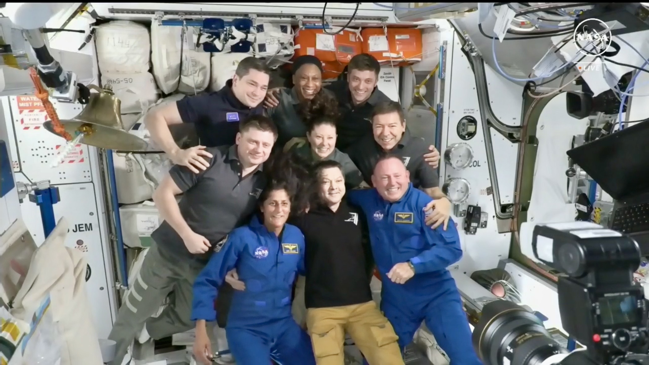 Nine astronauts float in a mostly white space station module, all smiling for a photo.