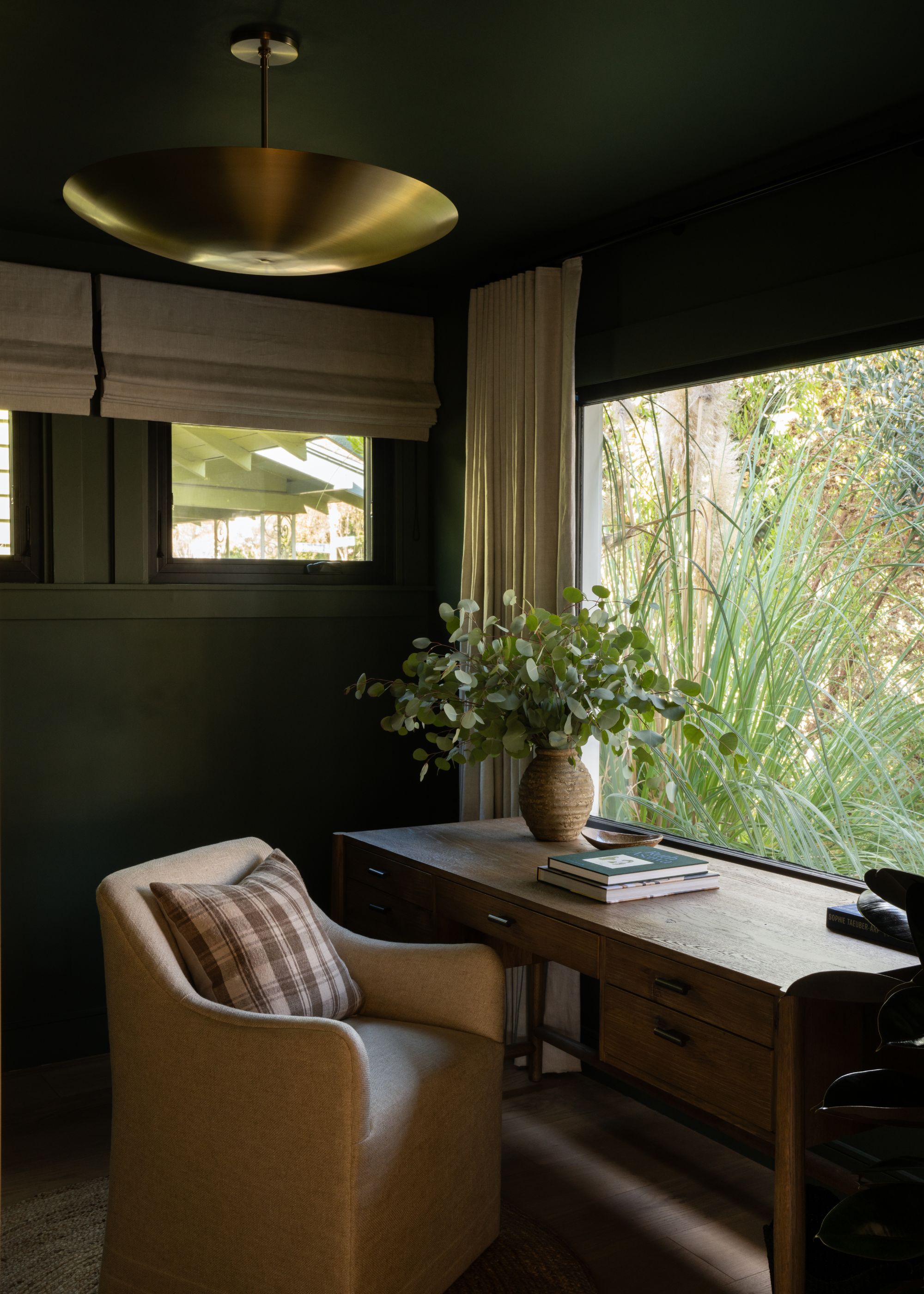 A home office with dark green color-drenched walls and ceiling, a large window with beige curtains, and a wooden desk in front looking out. A cream armchair and a plaid cushion.