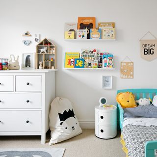 A white childrens bedroom with two bookshelves displaying colourful children's books, a white chest of drawers, and a turquoise bed with a lion cushion on it