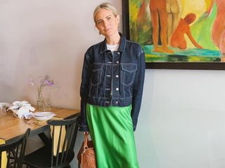 A woman wearing a dark-wash jean jacket buttoned up over a white T-shirt and a green slip skirt standing next to a table and in front of a painting