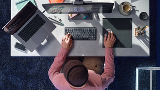 Et skrivebord set oppefra med en bærbar, en computerskærm og en mand, der skriver på et MX Mechanical tastatur