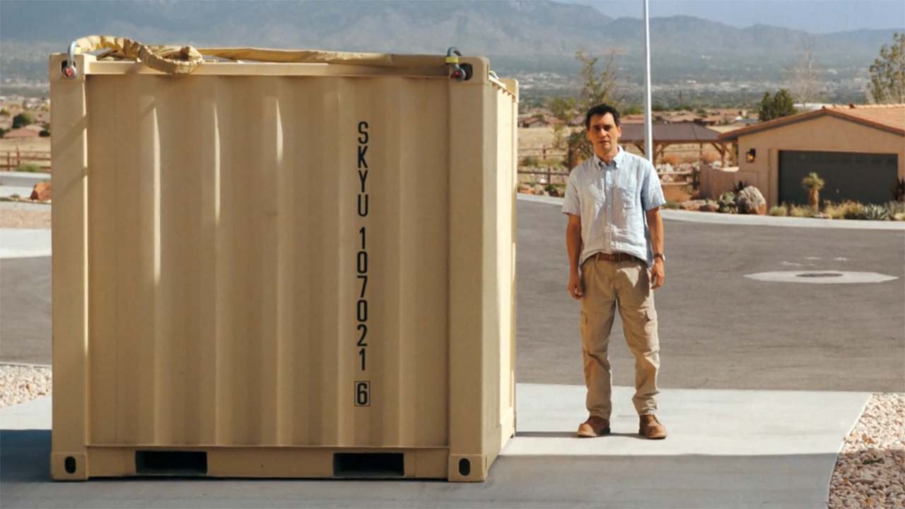 Manousos standing next to a giant, steel crate in Pluribus episode 9