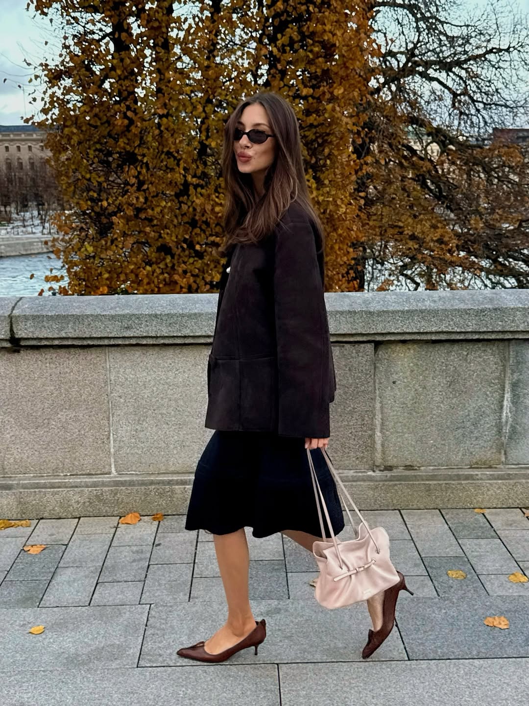 Felicia Akerstrom is photographed walking beside a river carrying the Prada Dada bag in pink wearing a dark jacket and skirt with Prada Antique pumps.