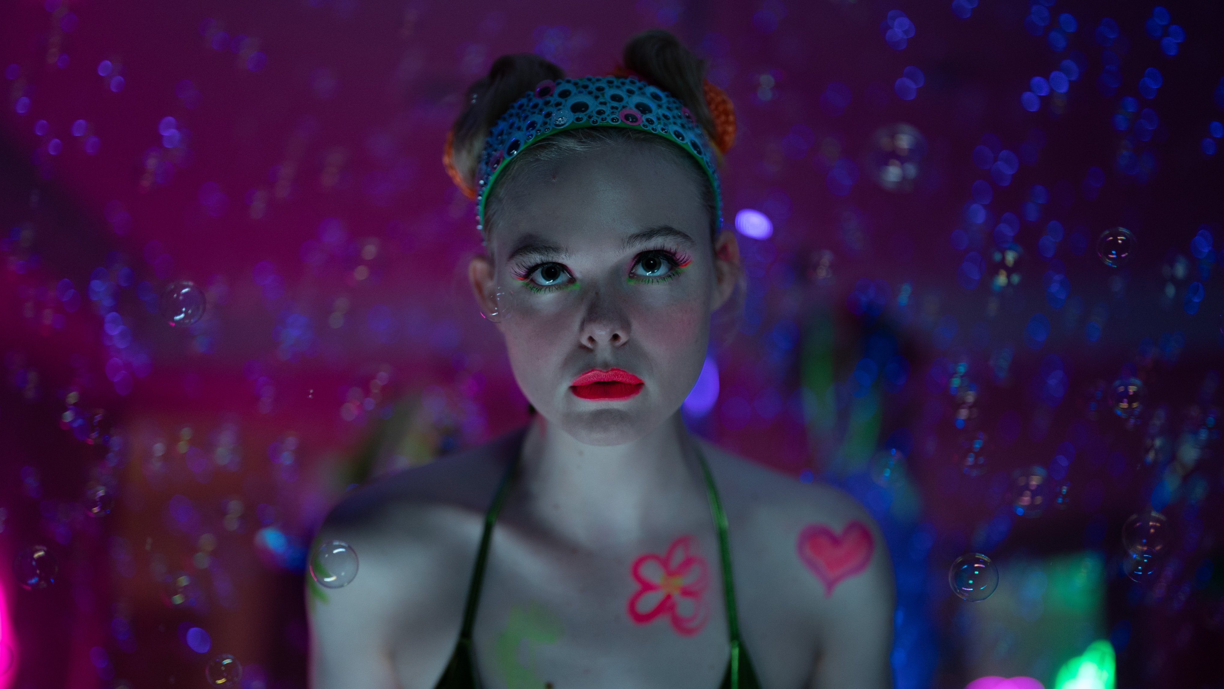 Elle Fanning as Margo wearing a tiara and neon pink lipstick with body paint. Bubbles surround her as she looks up at something off camera.