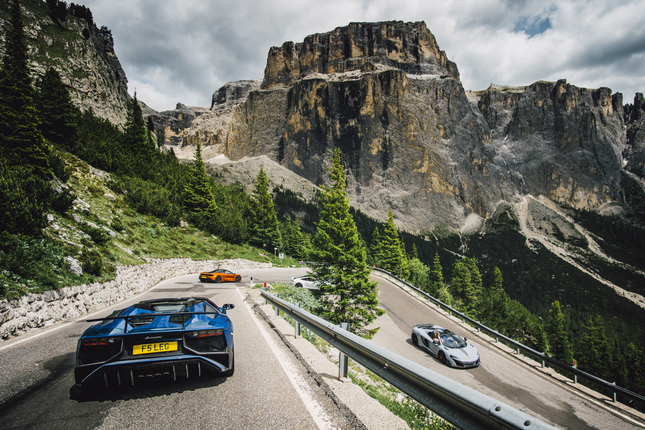 A line of supercars drives down a windy mountain road in italy