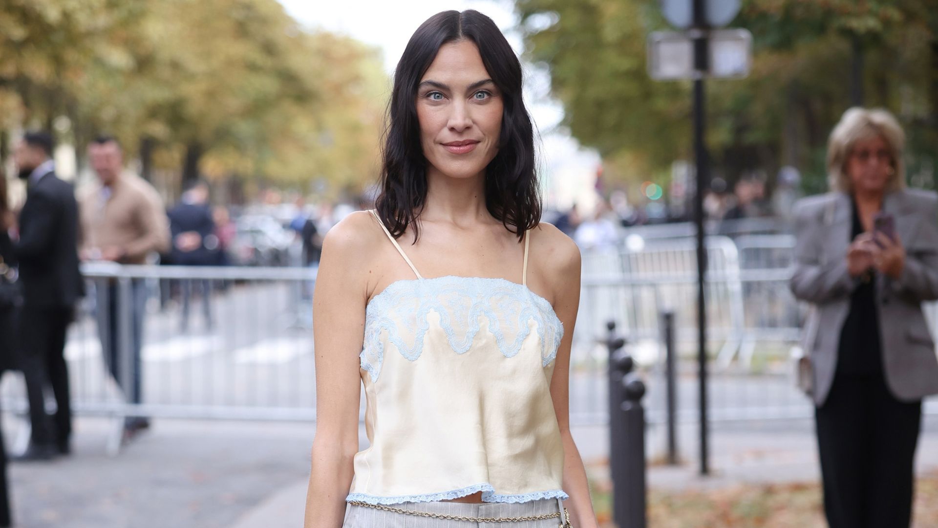 Alexa Chung poses for a photo outside of the Chloe SS27 runway show in Paris. She wears grey pinstripe trousers with a champagne lace-trimmed cami top and holds a beige Chloe Paddington bag.