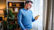 Mature man standing by the window at home and using a mobile phone