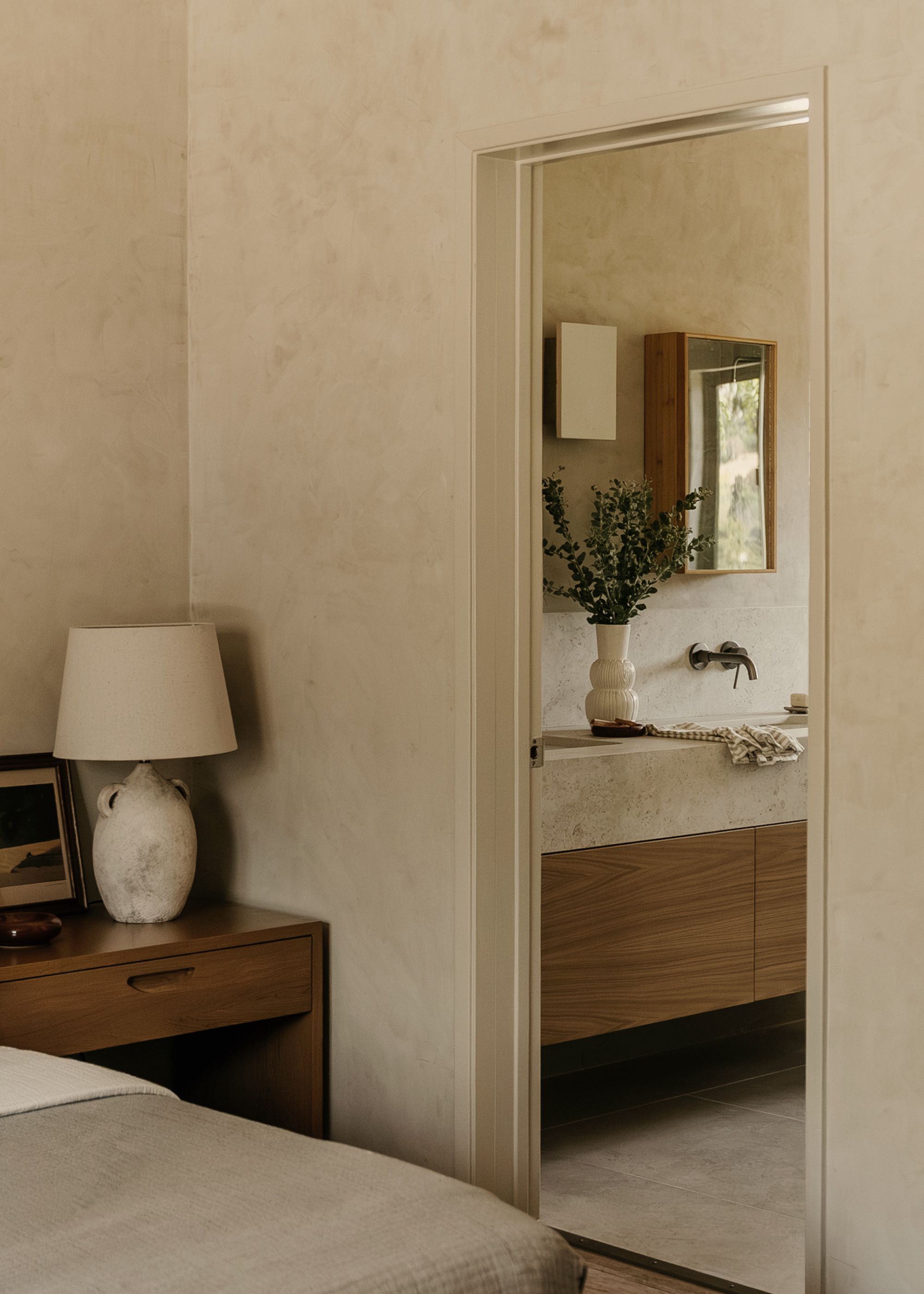 A minimalist Japanese bedroom with textured walls, a patinaed lamp, a vase of stems, and floating cabinets