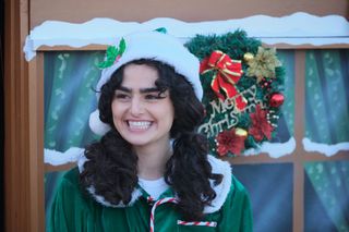 Zoe (Melina Sinadinou) stands outside Santa's grotto, dressed as an elf, smiling in a slightly forced way