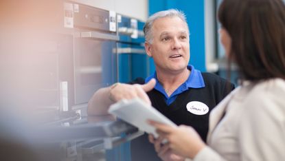An appliance expert speaks with a customer in front of a row of ovens.