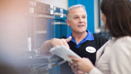 An appliance expert speaks with a customer in front of a row of ovens.
