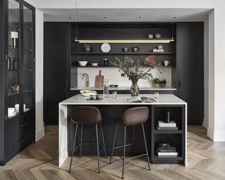 A kitchen with a black island with white wraparound counter top, seating and black floating shelves on the wall