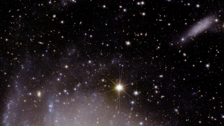 An image of the background of space with lots of bright white points of light. Toward the bottom of the image, there's a hazy purplish white glow gradually fading into the darkness as you get higher. Toward the top right, an edge-on object appears in the same color scheme as the glow.