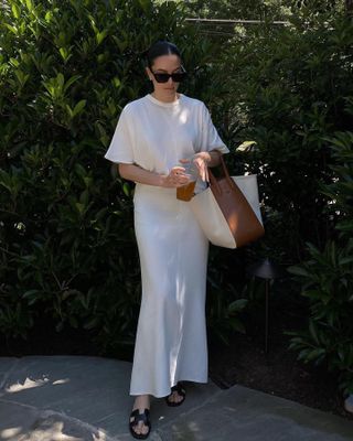 Atlanta-based style influencer Sarah Wisted poses outside next to bushes wearing oversize black sunglasses, an oversize white t-shirt, white silk slip skirt, two-tone tote bag, and black Hermès sandals.