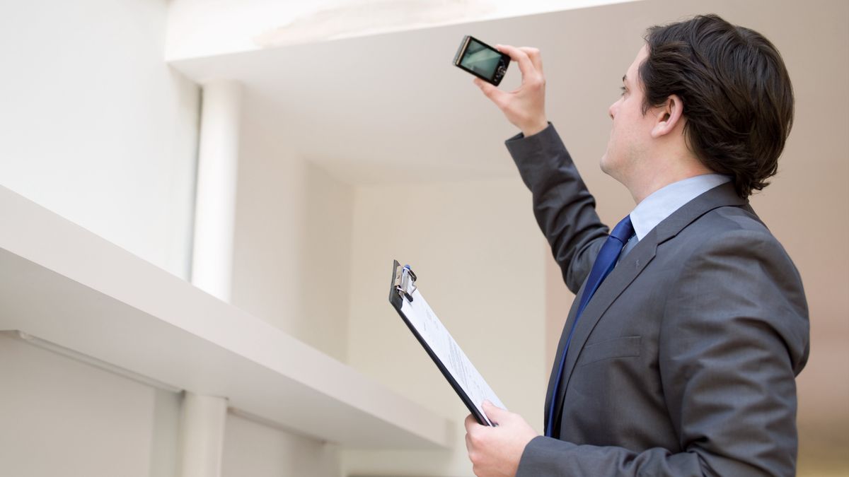 Surveyor in grey suit and blue tie with clipboard in hand standing in white painted room and photographing mark on ceiling with mobile phone