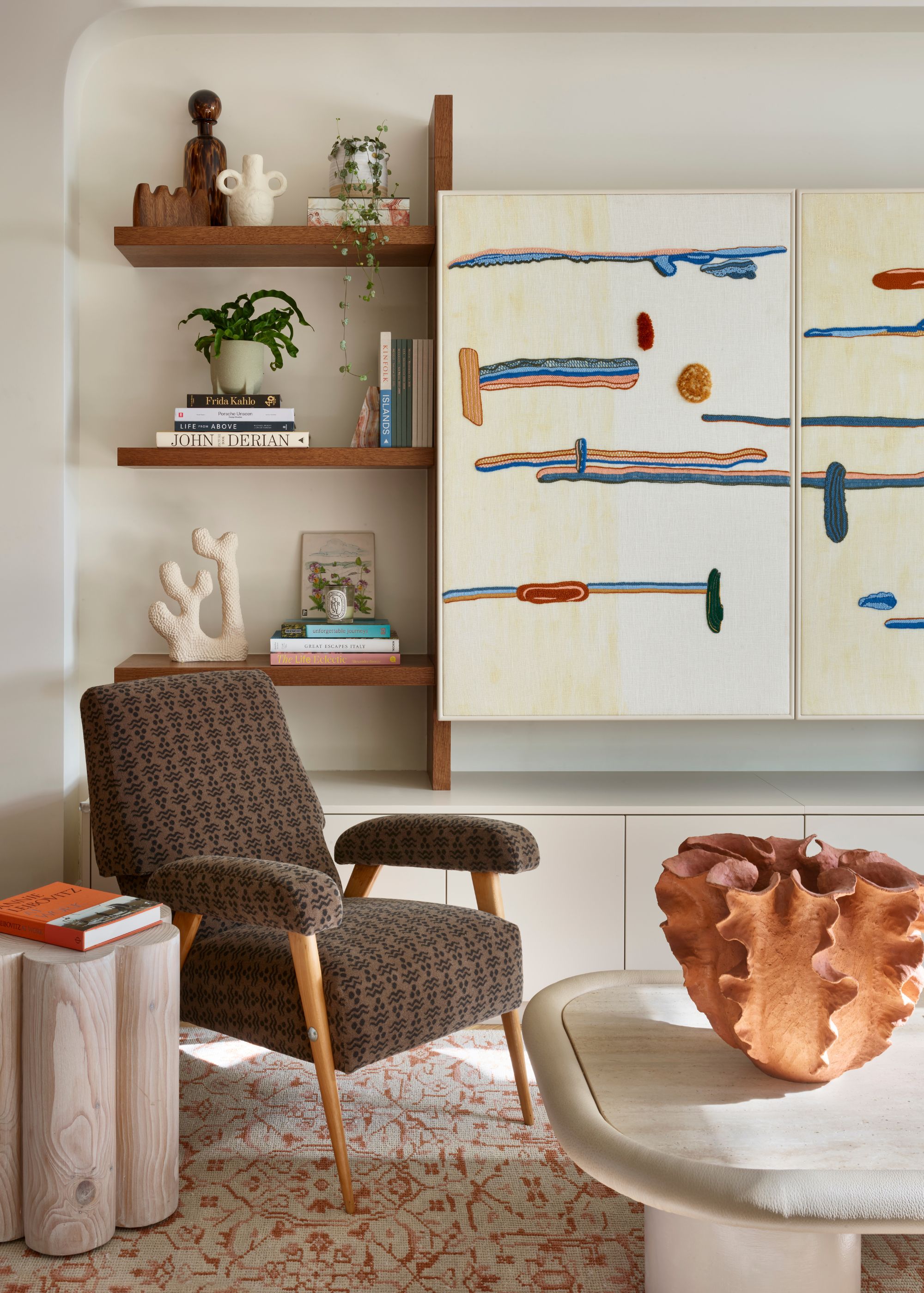 Corner of a neutral living room with wooden shelving and an abstract tapestry on the wall. A brown accent chair, a patterned rug, a sculptural coffee table, and a side table.