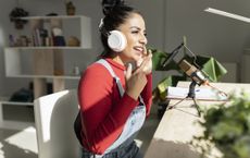 Woman sitting at home desk gestures while talking into microphone and wearing headphones recording a podcast