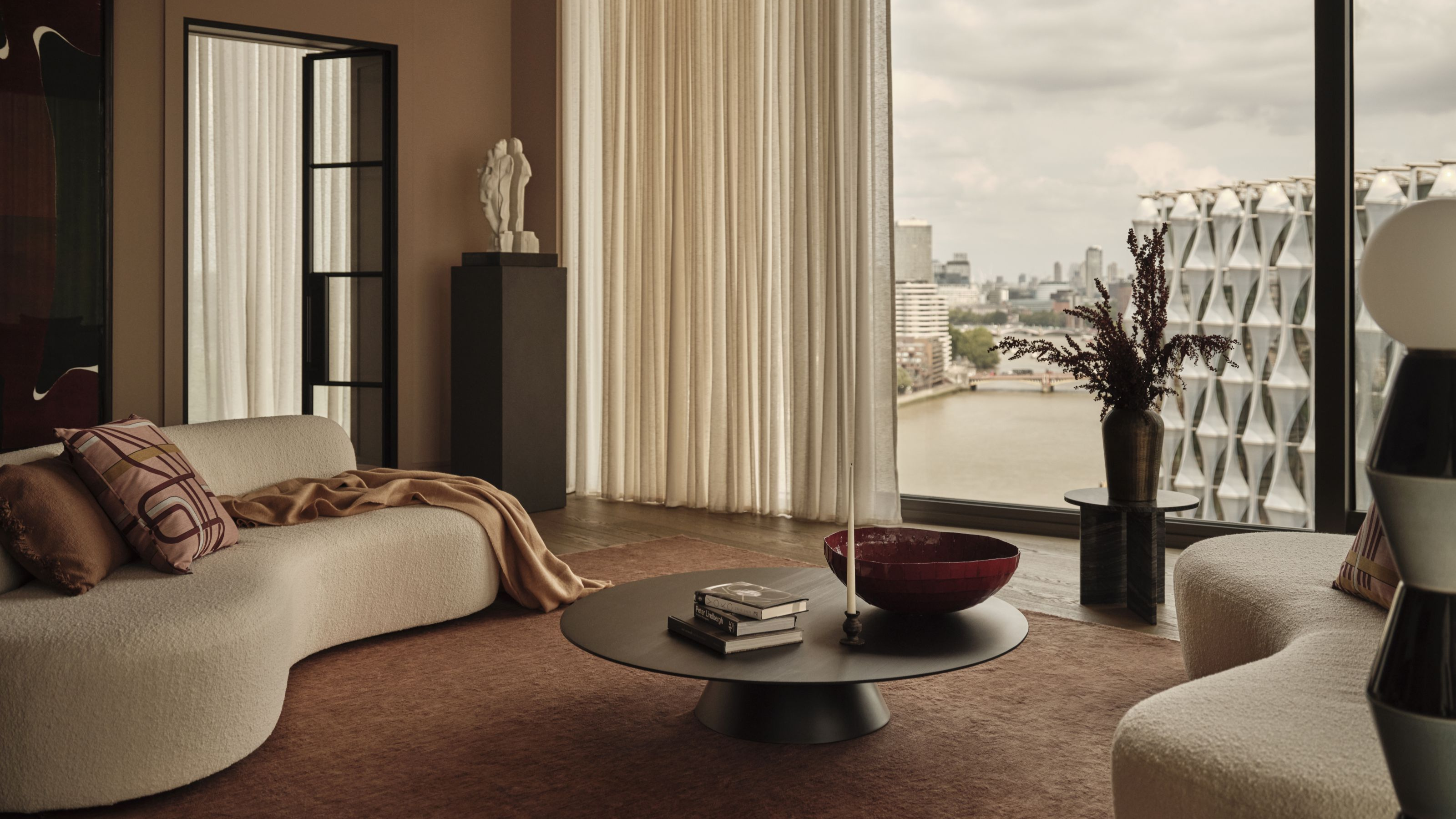 A modern living room with two white curved sofas facing each other with a round coffee table in the middle and full scale windows looking onto the London skyline with sheer cream curtains closed over a third of the window